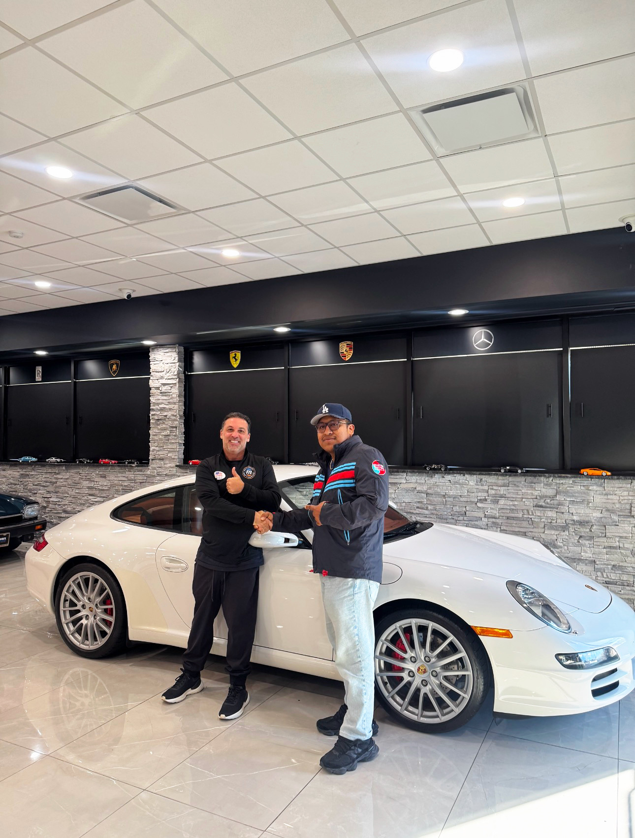Saúl shaking hands with a dealer beside a white Porsche 997 Carrera in a showroom