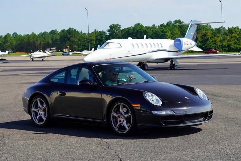 Black Porsche 911 Carrera parked on a tarmac in front of a private jet