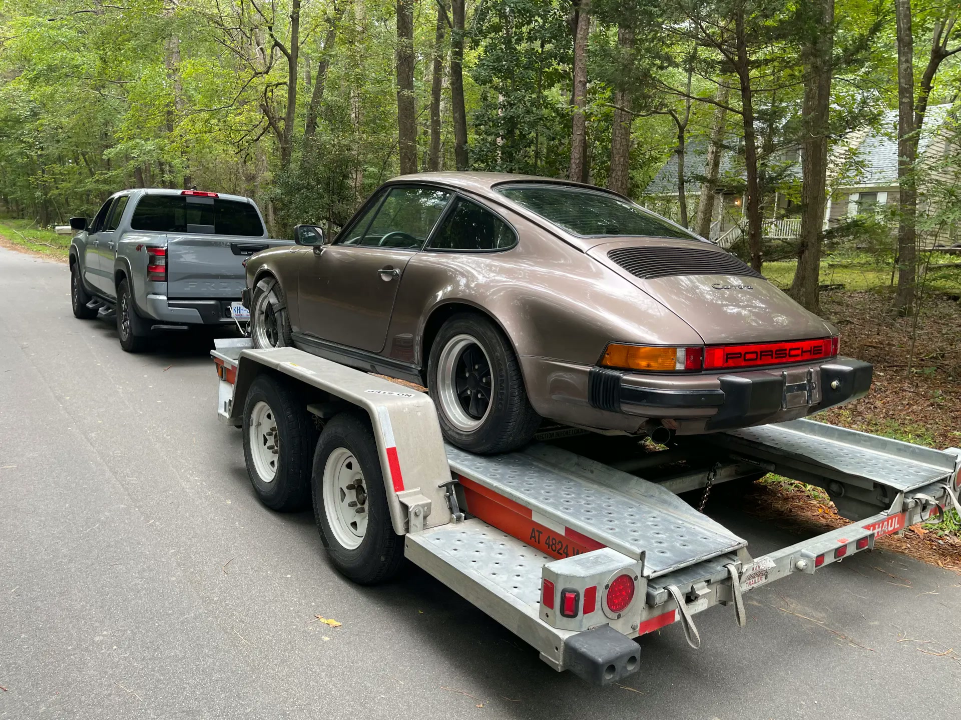 Brown Porsche 911 Carrera on a U-Haul trailer being towed by a gray pickup through a wooded North Carolina road