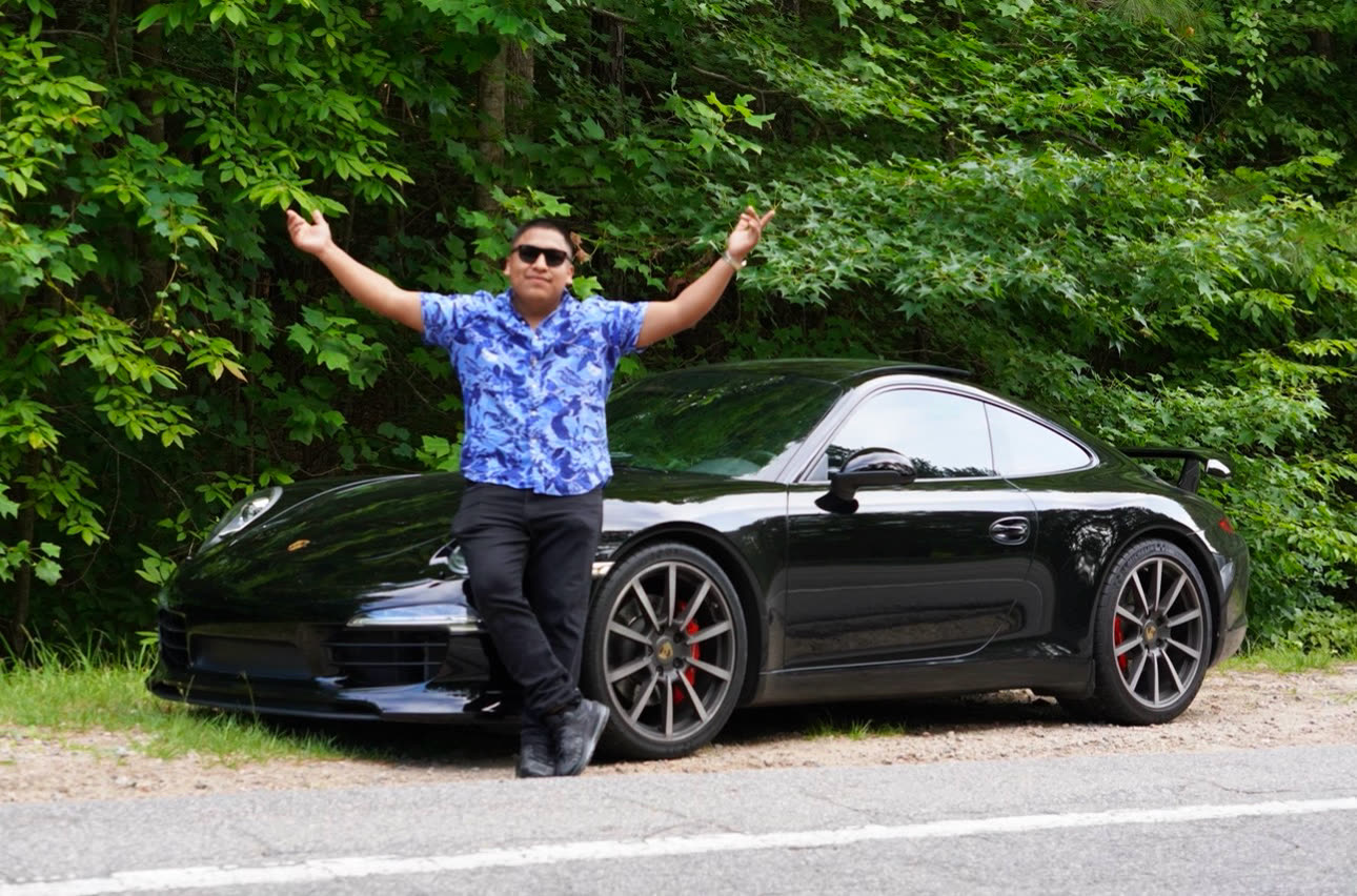 Saúl standing arms outstretched beside a black Porsche 911 cabriolet on a wooded road